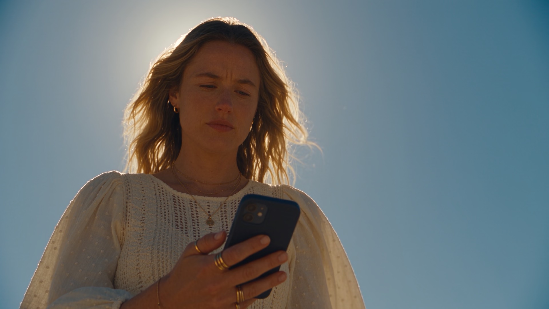 A mother looks down at her phone against a bright sky, thinking about what to send